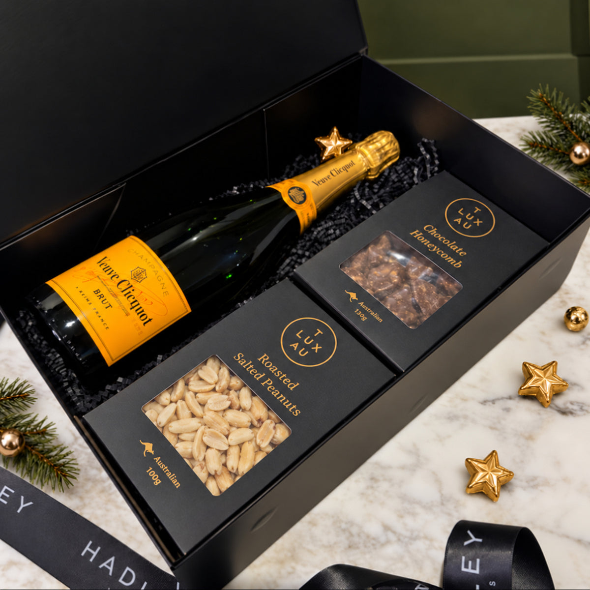 The Festive Champagne Indulger hamper with Veuve Clicquot Champagne, TLUX chocolate honeycomb and roasted salted peanuts in an open black gift box.