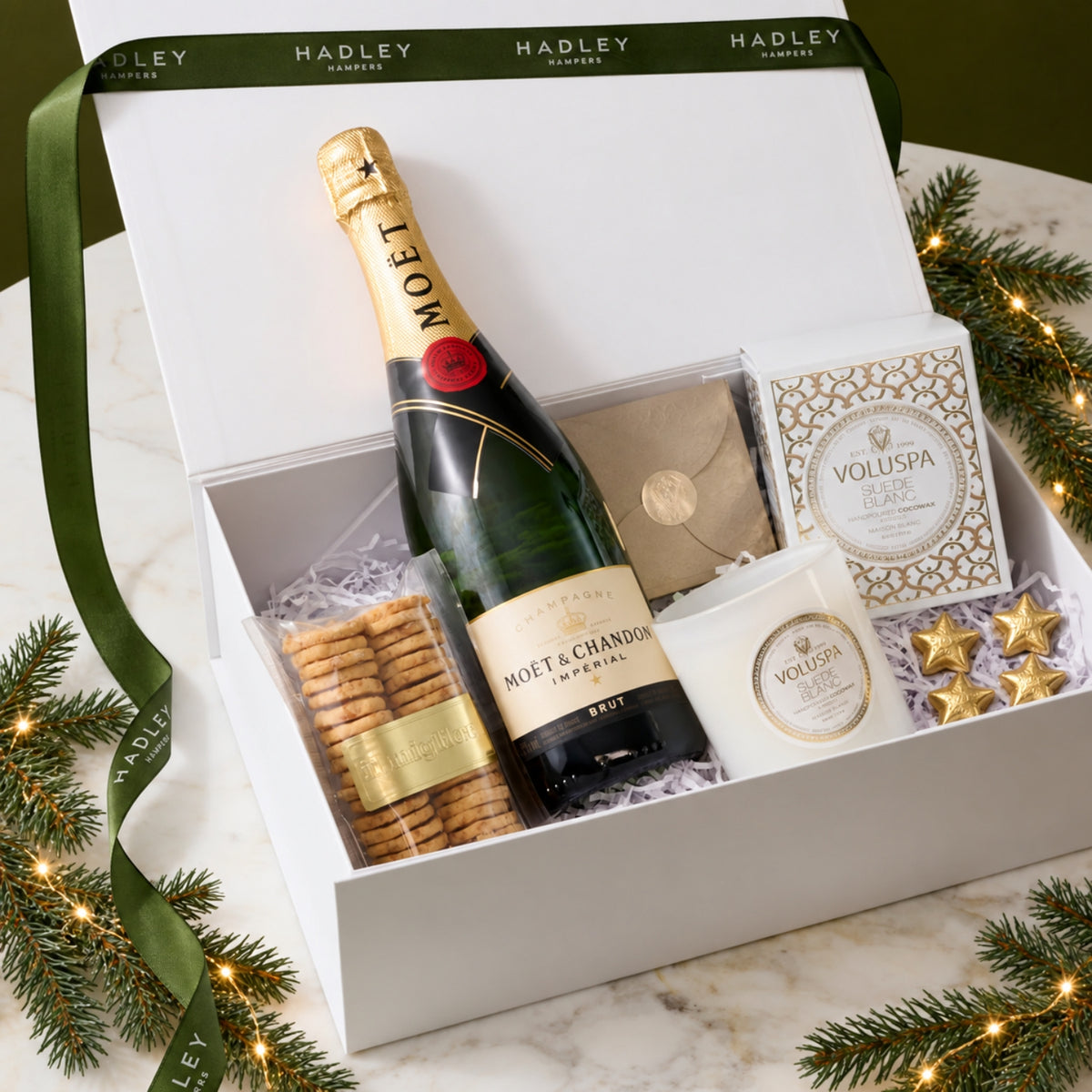 A luxury Christmas gift hamper from Hadley Hampers featuring a Moët & Chandon champagne bottle, Voluspa candle, chocolates, and savoury cheese bited, presented in a white gift box with a green ribbon, styled with pine branches and fairy lights on a marble surface.
