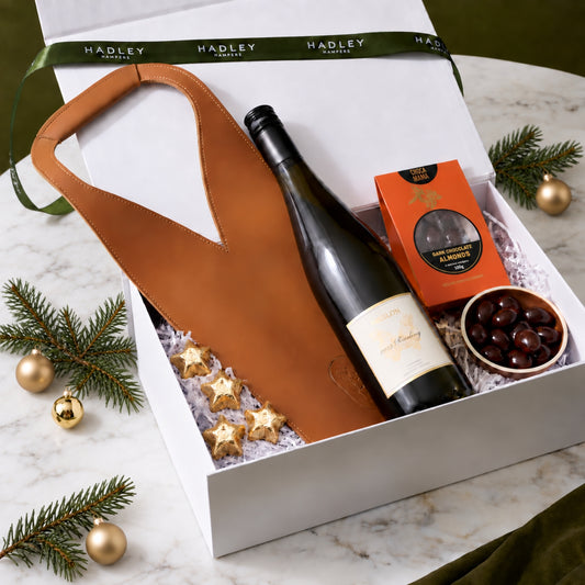 A luxury Christmas gift hamper from Hadley Hampers arranged in a white box on a marble surface. The gift set includes a brown leather wine carrier, a bottle of wine, an orange box of gourmet chocolates, and a bowl of dark chocolate almonds, surrounded by pine branches and gold ornaments.