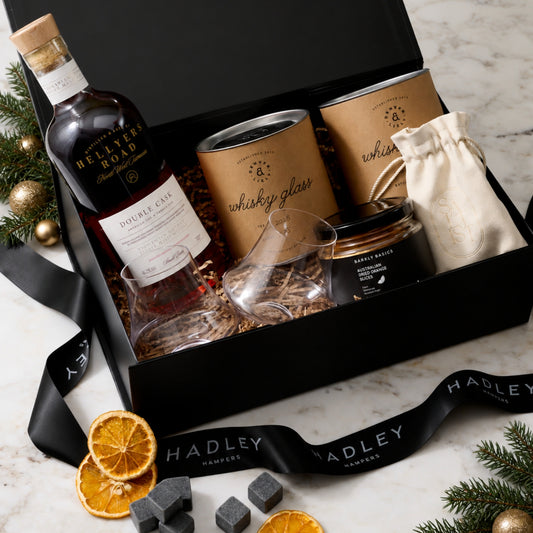 A curated gift box set from Hadley Hampers on a white marble surface. The black gift box contains a bottle of Hellyers Road Double Cask whisky, two premium whisky glasses, whisky stones, and dried oranges.