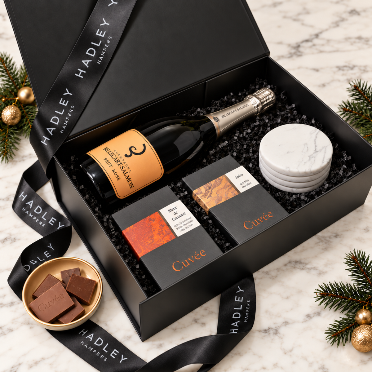Open black Christmas hamper with Billecart-Salmon rosé champagne, Cuvée Blanc de Caramel and Soleo chocolates, and four white marble coasters.