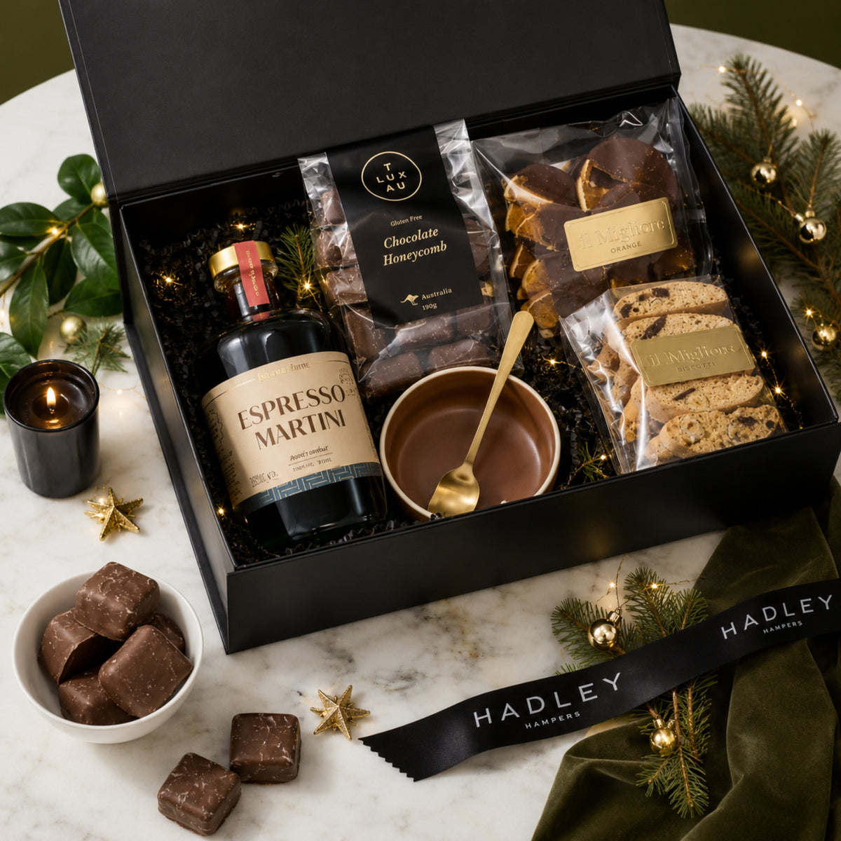 A luxury gift hamper from Hadley Hampers featuring a non-alcoholic Espresso Martini bottle, chocolate honeycomb, biscotti, and chocolates, arranged in a black box on a marble surface with holiday decorations.