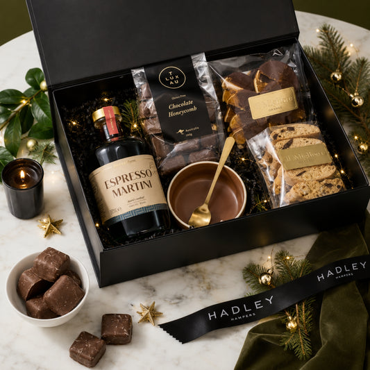 A luxury gift hamper from Hadley Hampers featuring a non-alcoholic Espresso Martini bottle, chocolate honeycomb, biscotti, and chocolates, arranged in a black box on a marble surface with holiday decorations.