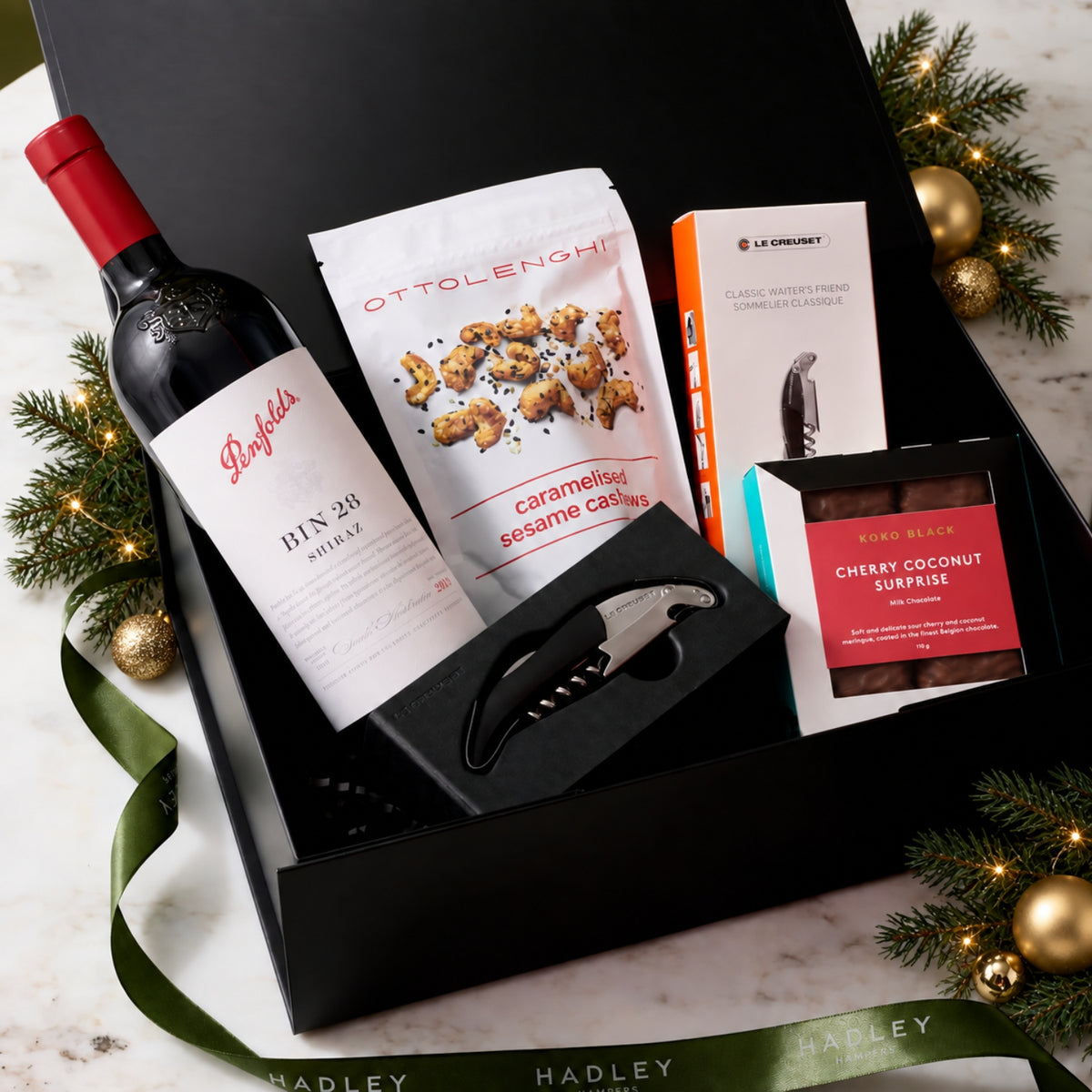 Festive Shiraz Sophisticate hamper with Penfolds Bin 28 Shiraz, Ottolenghi cashews, Koko Black Cherry Coconut Surprise chocolate and Le Creuset corkscrew in a black gift box with Christmas decorations.