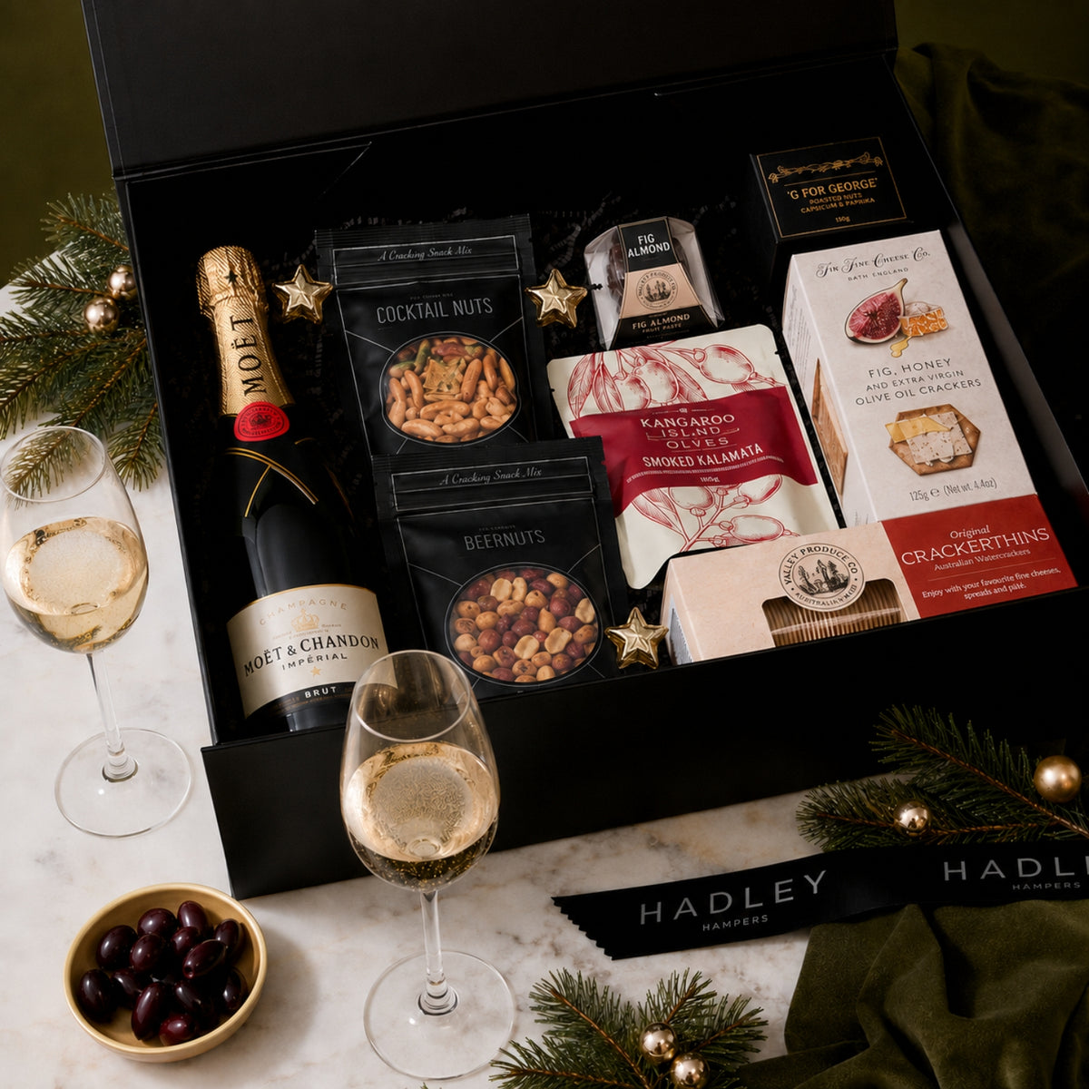 A luxury holiday gift hamper from Hadley Hampers, featuring a bottle of Moët & Chandon Brut, gourmet snacks, and champagne flutes, presented in a Christmas black gift box on a marble surface with festive pine branches and glasses of bubbly champagne.